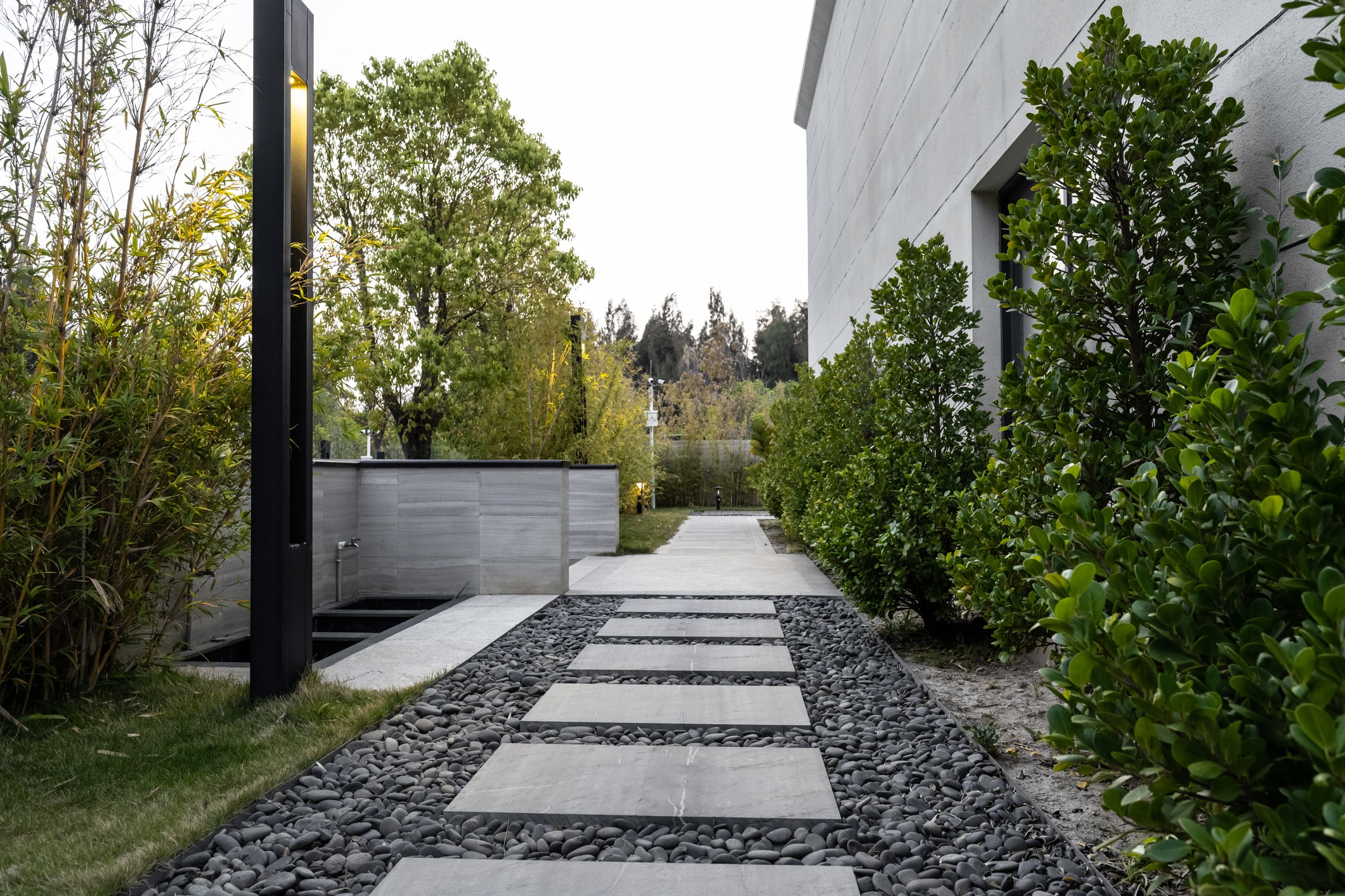 A modern garden pathway with rectangular stepping stones is flanked by lush greenery and pebbles, leading towards a landscaped area with trees and a lamp post.