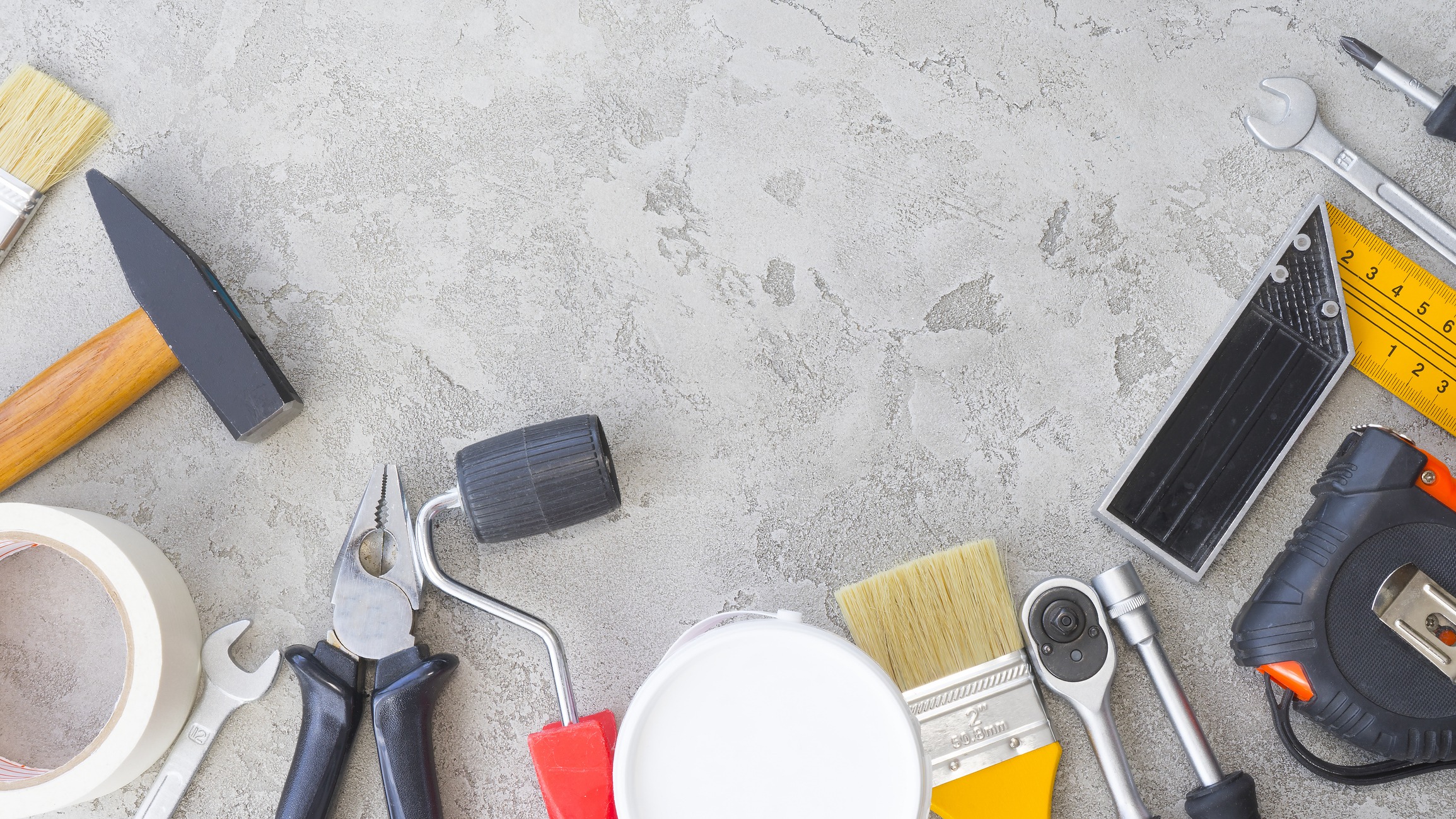 An assortment of tools and DIY equipment is spread out on a textured grey surface, including a hammer, tape measure, paintbrush, and other items.