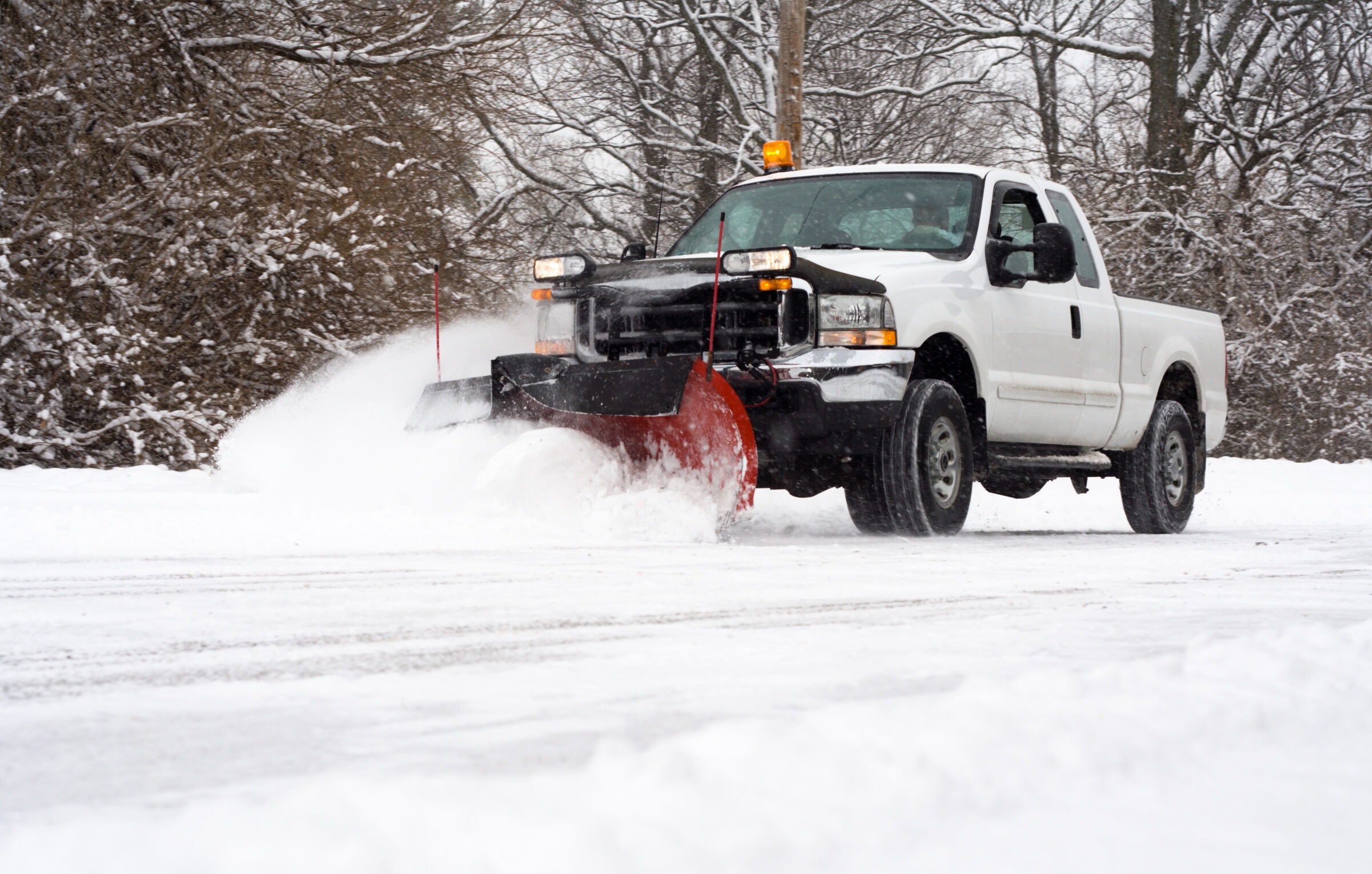 A white pickup truck equipped with a snow plow clears a road during a snowfall, with snow being pushed to the side and trees in the background.