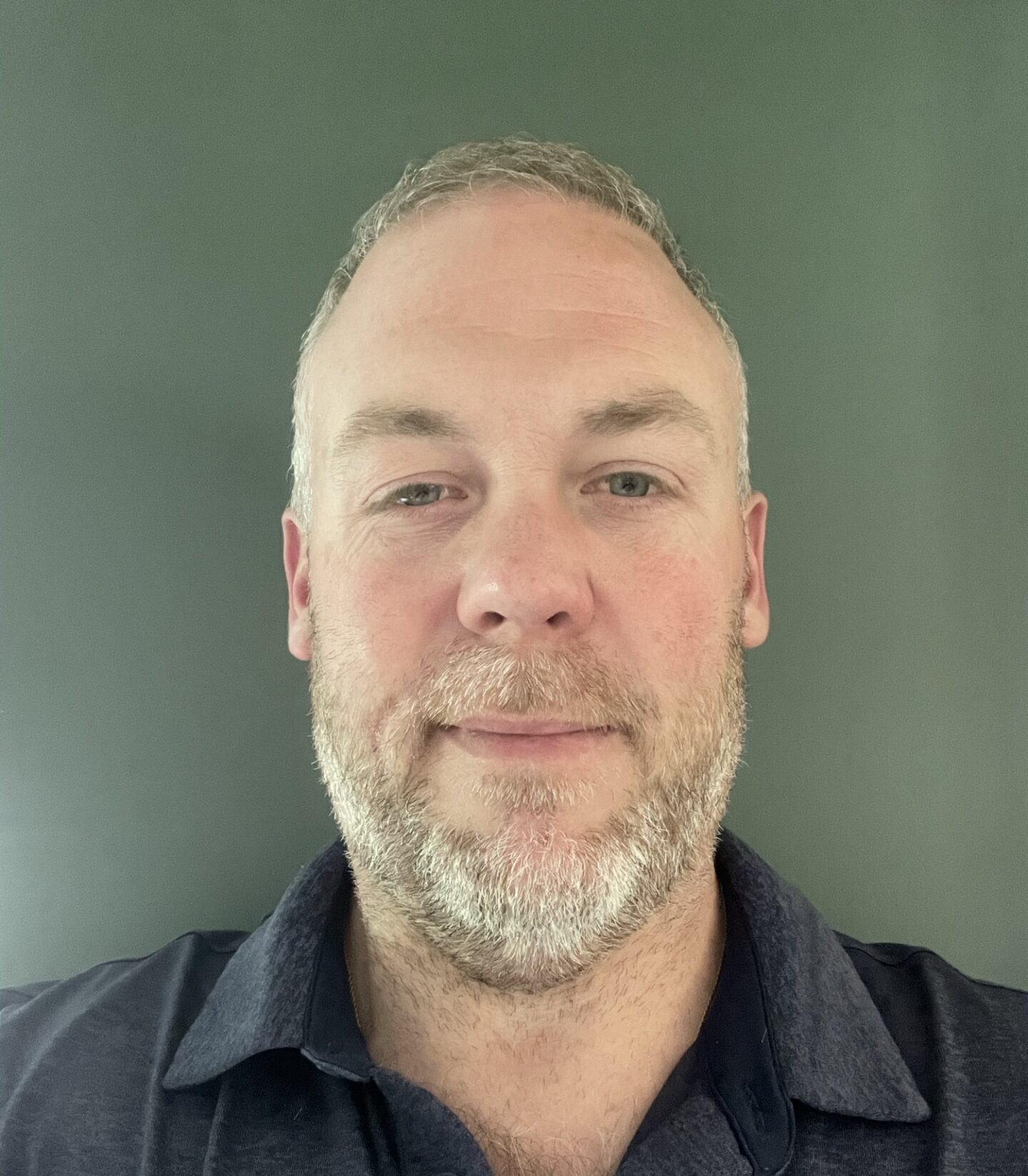 A person with short hair and facial stubble is smiling at the camera. They are wearing a dark collared shirt against a greenish-gray background.