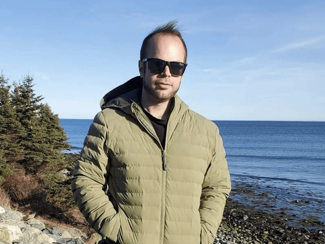 A person in sunglasses stands before a scenic coastline, wearing a green jacket. The ocean, trees, and rocky shore create a peaceful backdrop.