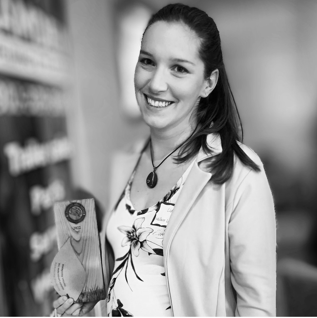 A smiling person holds an award in this black and white photo. They wear a blazer, a floral top, and a necklace, appearing happy and proud.