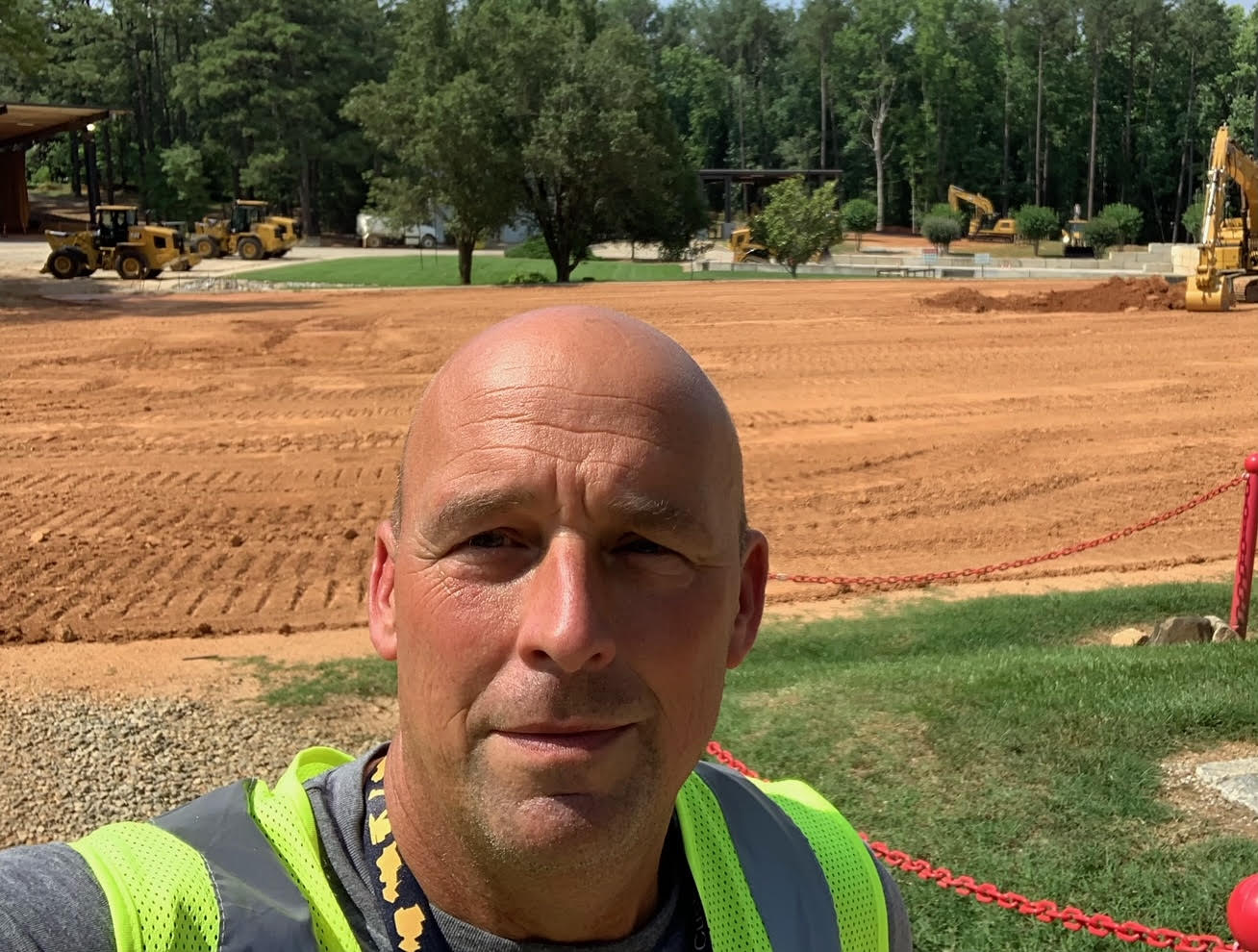 A person is taking a selfie at a construction site; they are wearing a high-visibility safety vest. In the background, there are excavators and bulldozers on dirt terrain.