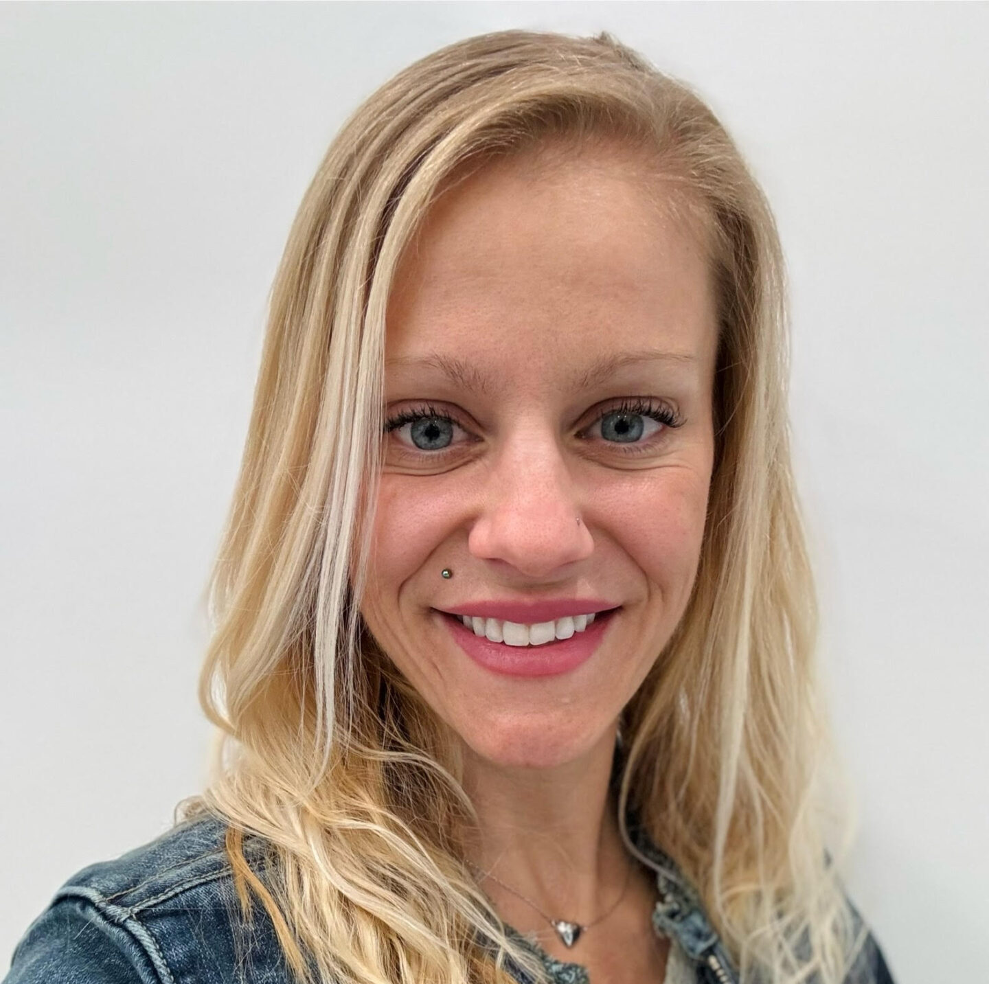 A smiling person with long blonde hair, blue eyes, and a nose piercing, wearing a denim jacket, against a plain white background.