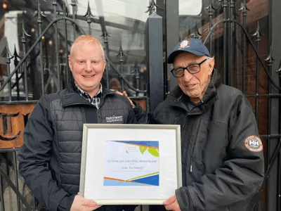 Two smiling people are holding a framed certificate with a graph and text, standing in front of a building with ornate metalwork. They appear pleased.