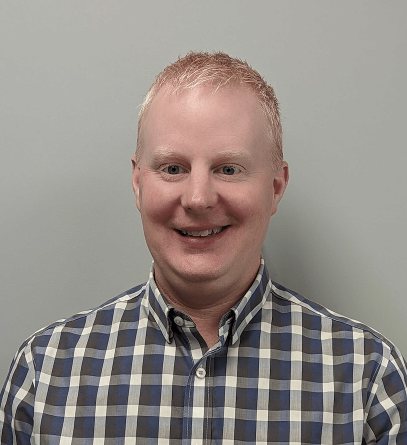 A person with short blond hair and a smiling expression, wearing a checkered shirt, is against a plain light-colored background.