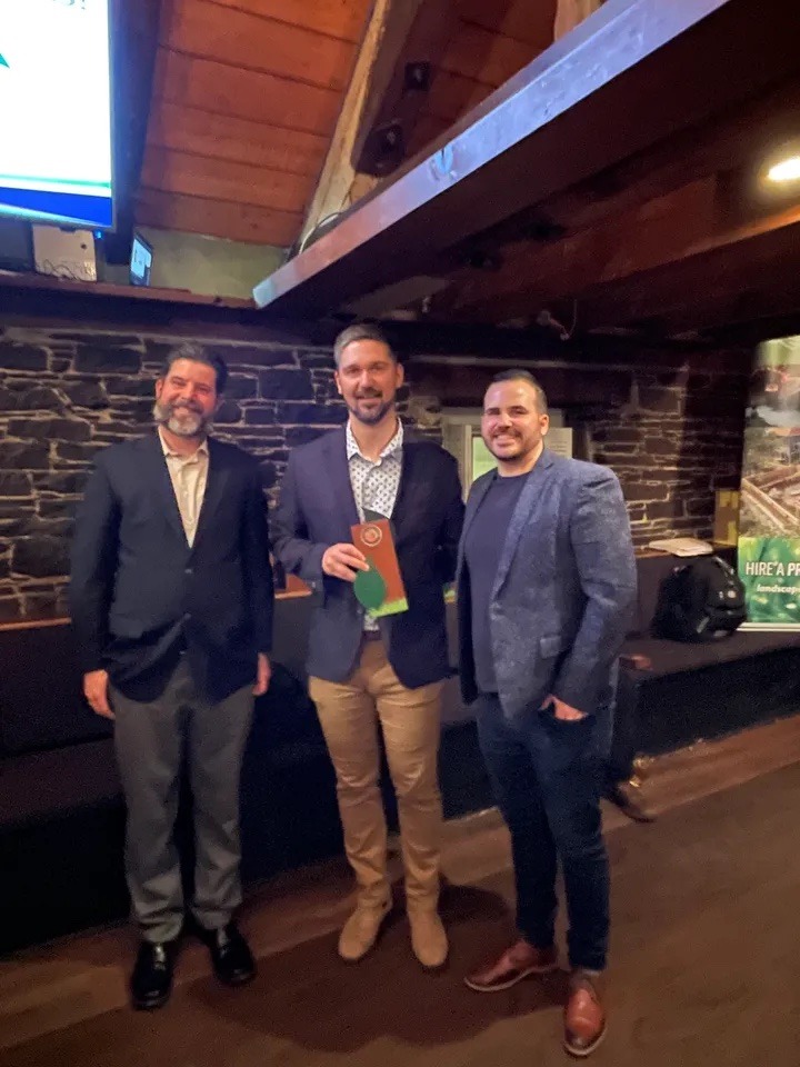 Three persons posing indoors with one holding an award. They dress semi-formally, standing in front of a stone wall and wooden beams.