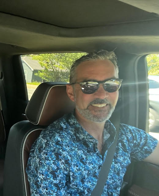 A smiling person with sunglasses is sitting in a car, wearing a blue patterned shirt, with a seatbelt on; residential area visible outside the window.