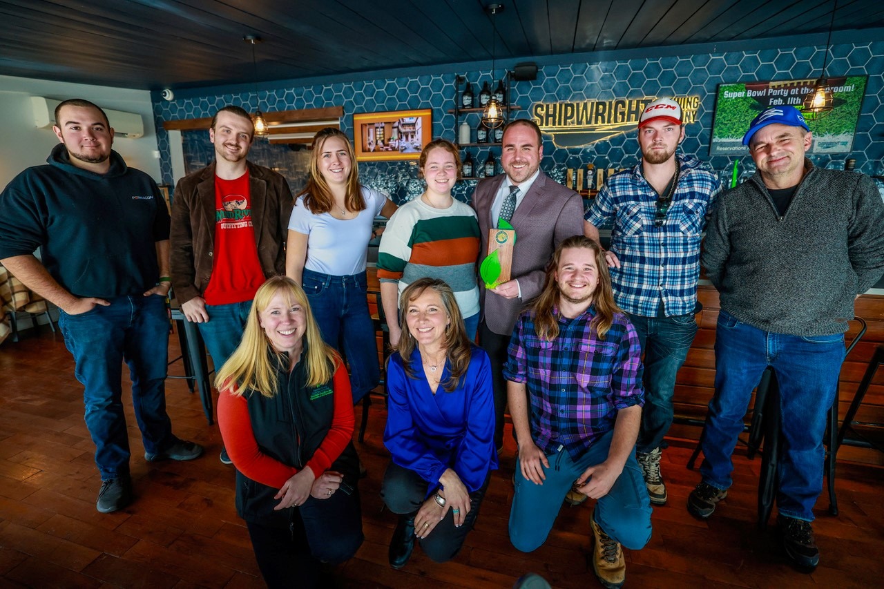 Nine individuals are posing for a group photo inside a room with a bar in the background. They are smiling and casually dressed.