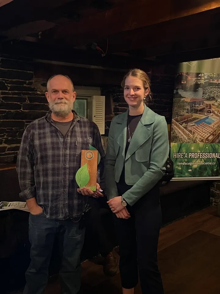 Two people are standing indoors with a wooden plaque, smiling for the photo. A banner reading "HIRE A PROFESSIONAL" is visible in the background.